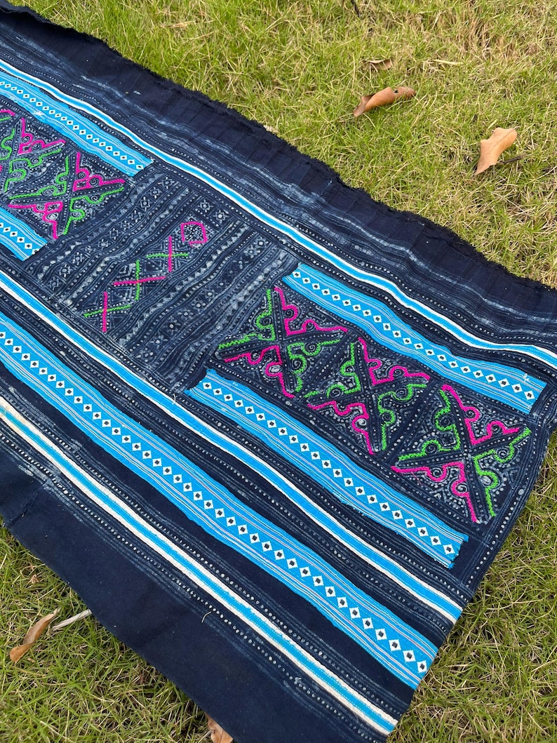 May include: A dark blue fabric with a white and blue striped pattern and intricate embroidery in pink, green, and white. The fabric has a frayed edge and is lying on a bed of green grass.