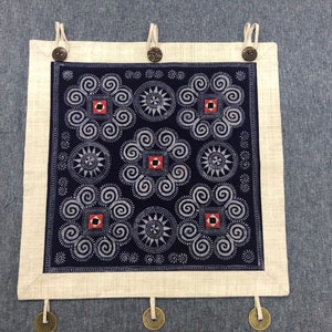 May include: A square wall hanging featuring a dark blue fabric panel with white spiral and star patterns, accented by small red and black squares. The panel is framed by a light beige border with three top loops secured by dark brown buttons.