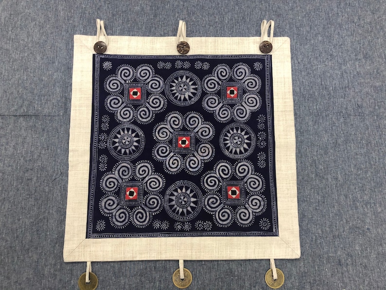 May include: A square wall hanging featuring a dark blue fabric panel with white spiral and star patterns, accented by small red and black squares. The panel is framed by a light beige border with three top loops secured by dark brown buttons.