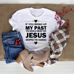 Może przedstawiać: Biała koszulka z napisem "If you bring up my past you should know that Jesus dropped the charges" w czarnych i złotych literach z sercami.
