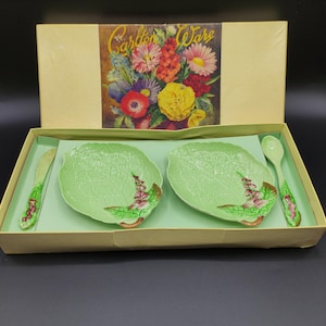 Könnte beinhalten: Ein Vintage-Carlton Ware Servierset in einer Box. Das Set enthält zwei blattförmige Teller, ein Messer und einen Löffel, alle in hellgrüner Farbe mit floralen Akzenten. Die Box hat eine Blumenillustration auf dem Deckel.
