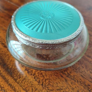 Puede incluir: Un frasco de vidrio redondo con una tapa circular que presenta un acabado de esmalte guilloché en color turquesa brillante. La tapa está bordeada por un borde de metal plateado decorativo con un patrón grabado intrincado sobre una base de vidrio.