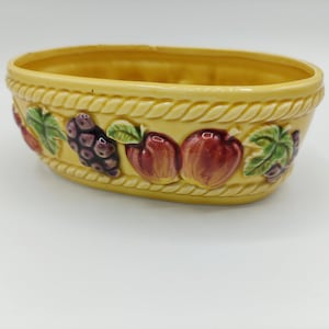May include: A yellow ceramic fruit bowl with a rope-like border. The bowl is oval-shaped and features embossed images of grapes, apples, and leaves in red, purple, and green. The bowl is empty.