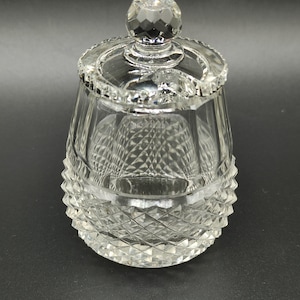 Könnte beinhalten: Ein klarer Glasbehälter mit Deckel. Der untere Teil des Behälters weist ein Diamantmuster auf, der obere Teil vertikale Linien. Der Deckel hat einen facettierten Knauf und einen dekorativen Rand.