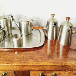 Op de afbeelding: Een thee- en koffieservies van roestvrij staal met een dienblad, twee theepotten, een melkkannetje en een suikerpot. Twee van de vaten hebben conische lichamen met houten handvatten en houten dekselknoppen, met een geborstelde metalen afwerking.