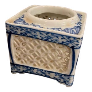 May include: A white ceramic incense burner with blue floral and geometric patterns. The square-shaped burner has a lattice design on the sides and a circular opening at the top. The edges are trimmed with blue detailing.