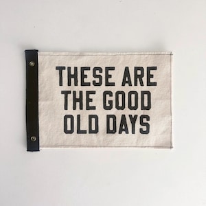 Könnte beinhalten: Ein rechteckiges Banner mit dem Text "THESE ARE THE GOOD OLD DAYS" in fetten schwarzen Buchstaben. Das Banner besteht aus einem hellbeigen Stoff mit einem schwarzen Rand auf der linken Seite und ist auf einer weißen Oberfläche montiert.