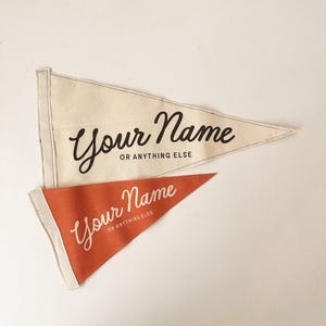 May include: Two pennant flags, one cream-colored and one burnt orange, with the text "Your Name OR ANYTHING ELSE" in a stylish script. The flags are triangular with a white border and are likely made of fabric.