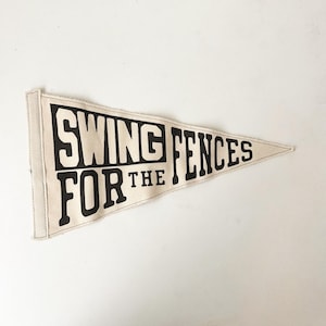 May include: A beige pennant flag with the text "SWING FOR THE FENCES" in bold black letters. The flag has a triangular shape and a stitched border. The pennant is made of a canvas-like material.