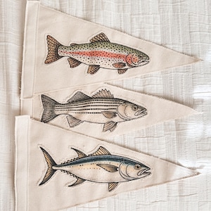 Op de afbeelding: Drie driehoekige wimpels met illustraties van vissen. De bovenste wimpel toont een regenboogforel met rode en groene markeringen. De middelste wimpel toont een gestreepte zeebaars. De onderste wimpel toont een tonijn. Allemaal op een crèmekleurige achtergrond.