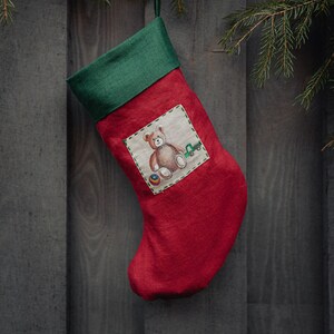 May include: Red Christmas stocking with a green cuff hanging on a wood panel wall. The stocking has a white patch with a brown teddy bear and a toy truck.