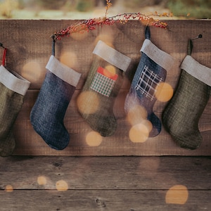 May include: Five Christmas stockings in shades of green and blue hang from a wooden wall. Each stocking has a neutral-colored cuff. One stocking has a red ribbon, and another has a gift-shaped patch. A sprig of berries is above.