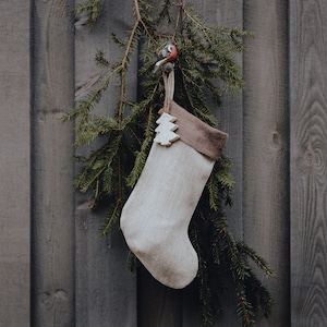 Pode incluir: Uma meia de Natal bege com punho castanho e um enfeite de árvore de madeira. Pendurada num ramo de pinheiro sobre um fundo de madeira envelhecida.