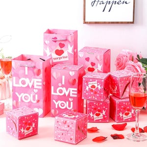 May include: A collection of pink gift bags and boxes decorated with hearts and doves. The bags feature the text "I Love You". Some boxes have the word "surprise". Two glasses of red liquid and rose petals are also visible.