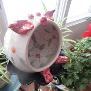 Puede incluir: Un macetero de cerámica con forma de cerdo con un esmalte blanco y un diseño floral. El macetero tiene forma de cerdo con un hocico y orejas rosas. El macetero está sobre un soporte negro.