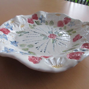 Puede incluir: Plato de servir de cerámica blanca con borde festoneado y diseño floral. El plato presenta flores rosas, azules y verdes, y un centro elevado con un patrón radiante.