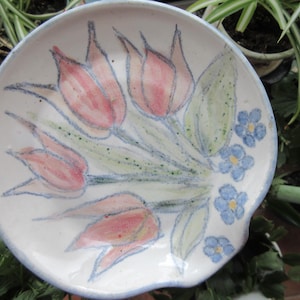 Puede incluir: Un plato de cerámica blanca con un diseño pintado a mano de tulipanes rosas y nomeolvides azules. El plato tiene un borde festoneado y un borde ligeramente elevado.