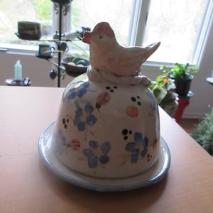 Puede incluir: Una mantequera de cerámica con tapa en forma de pájaro. El plato es blanco con diseños florales azules y mariquitas rosas. El pájaro es de color beige claro con detalles rosas.