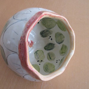 Puede incluir: Un cuenco de cerámica blanca con un borde rosa y hojas pintadas de verde y negro en el interior. El cuenco tiene una forma única, ligeramente irregular.