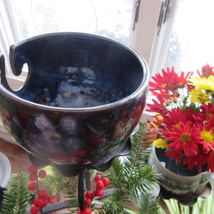 Puede incluir: Un cuenco de hilo de cerámica azul oscuro y marrón con un diseño curvo único. El cuenco está sostenido por un soporte de metal negro. Flores rojas y amarillas en el fondo. Ideal para sujetar el hilo mientras se teje o se hace ganchillo.
