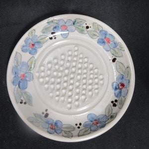 Puede incluir: Un rallador de ajo de cerámica blanca con un diseño floral azul y verde. El rallador tiene una superficie texturizada en relieve con pequeños bultos.