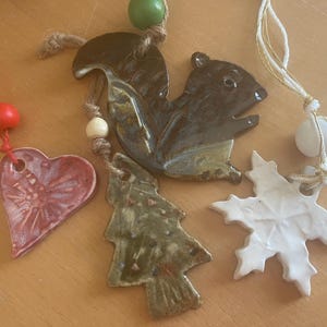 Puede incluir: Adornos de cerámica que incluyen un corazón rojo, una ardilla, un árbol de Navidad y un copo de nieve blanco. Cada adorno tiene una cuerda para colgar. Los adornos son en tonos de rojo, marrón, verde y blanco.