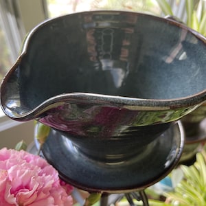 May include: A teal ceramic pour-over coffee maker with a brown rim. The handmade coffee maker has a wide, fluted rim and sits on a matching base. The pour-over is displayed with pink flowers and greenery. A stylish addition to any kitchen.