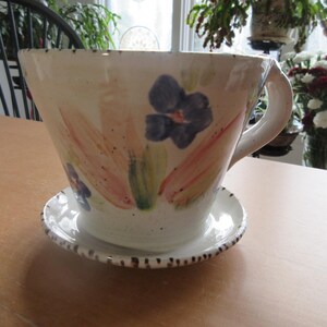 Puede incluir: Un macetero de cerámica blanca con forma de taza de té con asa. El macetero tiene un diseño floral con flores rosas, amarillas, verdes y azules. El macetero se asienta sobre un platillo a juego.