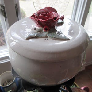 May include: White ceramic jar with a lid featuring a 3D red rose with green leaves. The jar has a smooth, glossy finish.