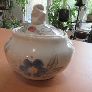 Puede incluir: Un azucarero de cerámica blanca con tapa. El cuenco presenta flores azules pintadas a mano con centros amarillos y hojas verdes. La tapa tiene un pequeño asa y detalles florales en rojo y azul. Un artículo de cocina decorativo.