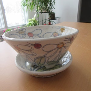 Puede incluir: Un cuenco de cerámica blanca con un diseño floral. El cuenco tiene un platillo a juego y está decorado con flores y abejas pintadas a mano en tonos azules, amarillos, rojos y verdes.