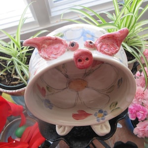 Puede incluir: Un cuenco de cerámica blanca con forma de cerdo con orejas y hocico rosas. El cuenco tiene un diseño floral con detalles en rosa, azul y amarillo. El cuenco está sobre una superficie negra.