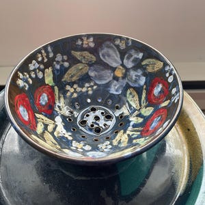 Puede incluir: Un cuenco de cerámica pintado a mano con un fondo azul oscuro, con diseños florales rojos, blancos y azules. El cuenco tiene una serie de agujeros en el centro y se asienta sobre un plato verde oscuro. Artículos de cocina decorativos.