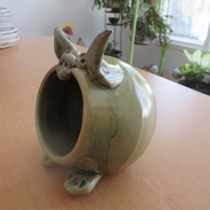 Puede incluir: Una alcancía de cerámica verde con una gran abertura en la parte posterior. La alcancía tiene una cara caprichosa con dos orejas y dos pies.