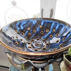 Puede incluir: Un colador de cerámica azul y marrón con agujeros para escurrir. El colador está sobre un soporte de metal negro.