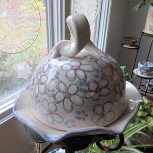 Puede incluir: Una fuente de cerámica con tapa abovedada, decorada con motivos florales blancos y azules. La fuente se apoya en un soporte decorativo. La tapa tiene un asa en forma de cisne. La fuente tiene aproximadamente 25 cm de diámetro.