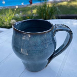 Puede incluir: Una taza de cerámica azul con cuerpo redondeado y asa. La taza tiene un esmalte azul oscuro con un sutil patrón moteado.