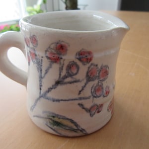 Puede incluir: Una jarra de crema de cerámica blanca con un diseño floral. La jarra tiene un asa y un pico. Las flores son rosas y rojas y están pintadas en la jarra.