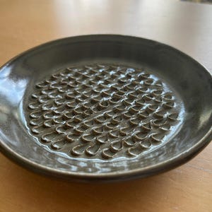 Puede incluir: Una placa ralladora de cerámica marrón oscuro con una superficie texturizada. La placa tiene un diseño poco profundo y redondo con un borde elevado. La superficie de rallado está cubierta de pequeñas crestas en forma de lágrima, muy juntas.