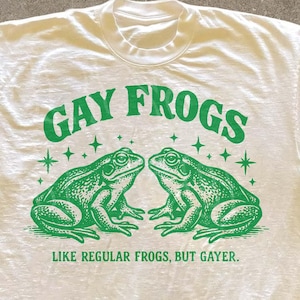 Może przedstawiać: Kremowa koszulka z zieloną grafiką przedstawiającą dwie żaby i napisem "GAY FROGS". Poniżej znajduje się napis "LIKE REGULAR FROGS, BUT GAYER." Koszulka ma okrągły dekolt i krótkie rękawy.