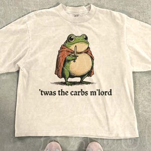 May include: A natural-colored short-sleeve t-shirt featuring a graphic of a round green frog wearing a red cape and holding a small sword. Below the frog, the text reads 'twas the carbs m'lord in a dark font. The shirt has a crew neckline.