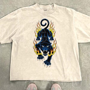 A white t-shirt with a graphic design of a black panther with flames on its back, and a pair of white sneakers with black stripes.
