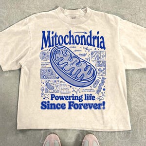 May include: A beige short-sleeve t-shirt featuring a large blue graphic of a mitochondrion in the center. The text reads "Mitochondria" at the top and "Powering life Since Forever!" at the bottom, surrounded by small scientific diagrams.
