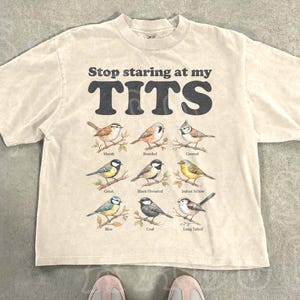 Puede incluir: Camiseta beige con el texto negro "Stop staring at my TITS". Debajo, ilustraciones de varios pájaros. La camiseta está colocada sobre una superficie de hormigón.