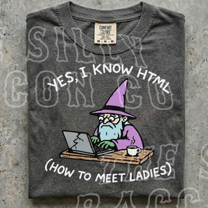 Könnte beinhalten: Ein dunkelgraues T-Shirt mit einer Grafik eines Zauberers mit violettem Hut und Bart, der an einem Schreibtisch mit einer Kaffeetasse an einem Laptop tippt. Der Text lautet: YES, I KNOW HTML (HOW TO MEET LADIES). Das Shirt hat ein Comfort Colors Etikett.