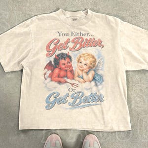 Puede incluir: Una camiseta beige de manga corta con un estampado gráfico de un diablo rojo y un ángel rubio. El texto dice: You Either... Get Bitter, Or Get Better. La camiseta tiene cuello redondo y un corte relajado, mostrada con pantalones vaqueros azul claro.