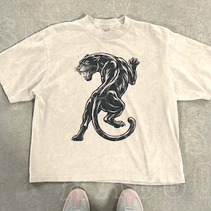 Pode incluir: Uma camiseta de cor natural com um gráfico de pantera negra. A camiseta tem gola redonda e mangas curtas. O texto "THE SILLY RACCOON CO" está impresso no canto superior esquerdo. A palavra "natural" está impressa no canto inferior direito.