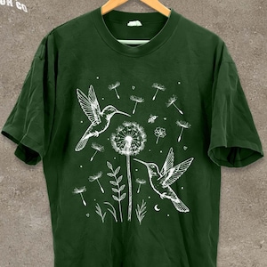 A green t-shirt with a white illustration of two hummingbirds hovering over a dandelion surrounded by flowers and plants.