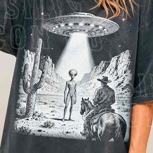 A woman wearing a black t-shirt with a graphic design depicting an alien spacecraft hovering over a desert landscape with a cowboy on horseback.