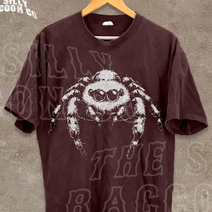 A dark brown t-shirt with a white spider graphic printed on it, hanging on a wooden hanger against a concrete wall.